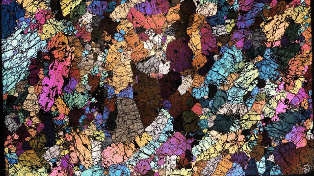 Ein polarisationsmikroskopisches Bild zeigt eine Vielzahl von bunten, unregelmäßig geformten Strukturen, die an ein Mosaik erinnern. Es stellt die mikroskopische Struktur eines Gesteins dar.
