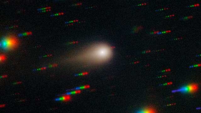 Ein astronomisches Bild zeigt einen Kometen mit einem leuchtenden Kern und einem diffusen Schweif, der sich nach rechts erstreckt. Um den Kometen herum sind bunte, spektrale Streifen zu sehen, die auf die Bewegung von Sternen oder anderen Himmelskörpern hinweisen. Der Hintergrund ist dunkel, was den Kontrast zu den hellen, farbigen Streifen und dem Kometen verstärkt. Das Bild vermittelt den Eindruck von Bewegung und Dynamik im Weltraum.
