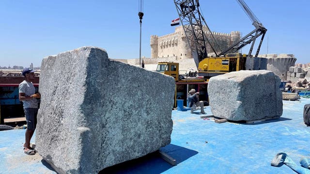Ein Arbeiter steht neben einem großen, rechteckigen Steinblock an einer Stelle mit blauem Bodenbelag. Im Hintergrund ist ein gelber Kran zu sehen, der einen weiteren Steinblock hebt. Eine historische Festung erhebt sich im Hintergrund unter einem klaren blauen Himmel. Eine ägyptische Flagge weht auf dem Kran. Die Szene vermittelt den Eindruck von archäologischen Arbeiten in der Nähe eines historischen Ortes.