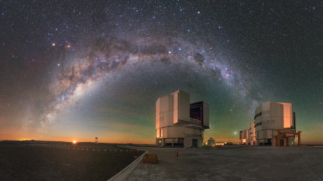 Sternenklare Nacht über dem Paranal-Observatorium in Chile. Der Bogen der Milchstraße erstreckt sich über den Himmel, während zwei große Teleskope im Vordergrund stehen. Der Horizont zeigt einen sanften orangefarbenen Schein, der auf den Sonnenuntergang oder die Dämmerung hindeutet.