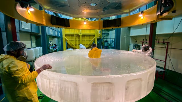 Drei Personen in Schutzkleidung und Atemmasken arbeiten in einem Labor an einem großen, runden, transparenten Objekt. Es ist der Hauptspiegel des 3,6-Meter-Teleskops der Europäischen Südsternwarte ESO in Chile, der hier gerade einer Reinigung unterzogen wird. Die Umgebung ist industriell, mit gelben und grünen Elementen. Oben hängt eine große, runde, gelbe Struktur mit Lichtern.