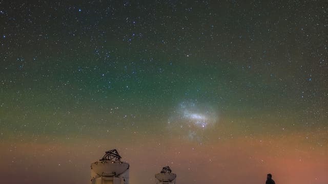 Ein nächtliches astronomisches Bild zeigt einen klaren Sternenhimmel mit zwei markanten Galaxien, der Großen und der Kleinen Magellanschen Wolke, die über einem Observatorium leuchten. Im Vordergrund sind mehrere Teleskope auf Schienen zu sehen, die auf einer Plattform stehen. Eine Person steht rechts und blickt in den Himmel. Der Himmel zeigt einen sanften Farbverlauf von Grün bis Orange, was auf atmosphärische Effekte hinweist.