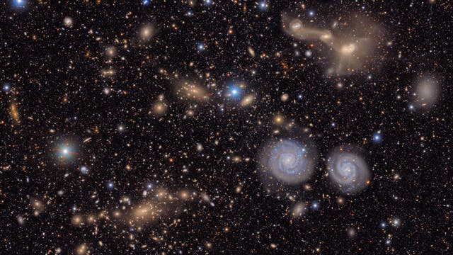 Eine dichte Ansammlung von Galaxien im Weltraum, darunter zwei markante Spiralgalaxien in der unteren rechten Bildhälfte. Der Hintergrund ist mit zahlreichen Sternen und weiteren, teils verschwommenen Galaxien gefüllt. Die Szene zeigt die Vielfalt und Weite des Universums.