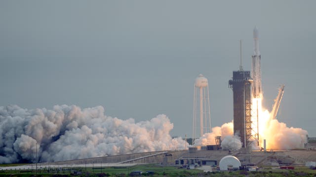 Die Rakete Falcon Heavy hebt vom Raumfahrtbahnhof  Kennedy Space Center in Florida ab. Der Himmel ist grau, der Raketentreibstoff verbrennt orange, links im Bild sieht man Wolken des verbrannten Treibstoffs