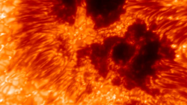 Nahaufnahme einer Sonnenoberfläche mit dunklen Sonnenflecken und leuchtenden, wirbelnden Plasmamustern in Rot- und Orangetönen. Die Textur zeigt die dynamische Aktivität der Sonne.