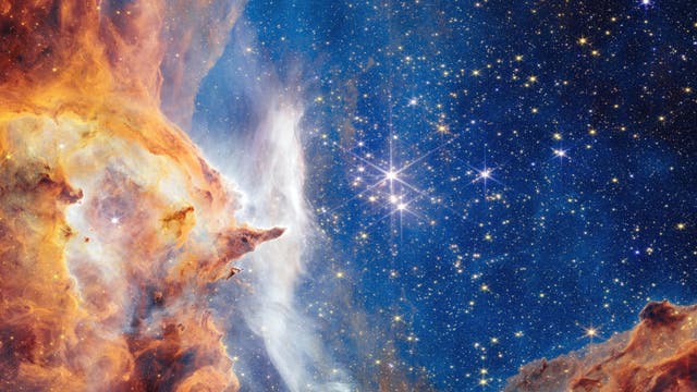 Ein farbenfrohes astronomisches Bild zeigt eine dichte Ansammlung von Sternen vor einem tiefblauen Hintergrund. Links im Bild sind leuchtende orangefarbene Gas- und Staubwolken zu sehen, die sich in den Weltraum erstrecken. Die Wolken haben eine komplexe, strukturierte Form, die an eine Nebelregion erinnert. Rechts im Bild funkeln zahlreiche Sterne, einige davon besonders hell, und bilden ein beeindruckendes Sternenfeld. Das Bild vermittelt die Weite und Schönheit des Universums.