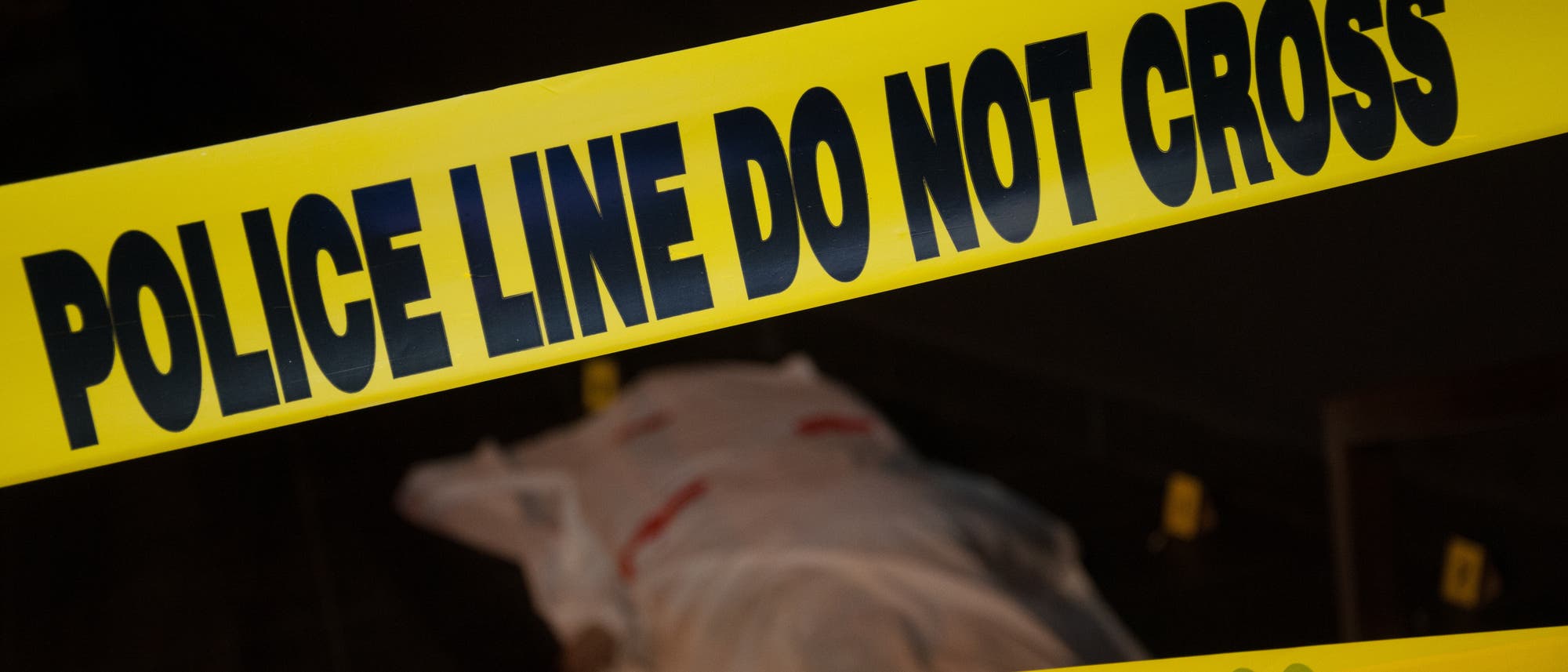 (amerikanisches) Polizei-Absperrband, dahinter schemenhaft eine 'Leiche' (vermutlich eine Schaufensterpuppe unter einem rot beschmierten weißen Laken). (amerikanisches) Polizei-Absperrband, dahinter schemenhaft eine 'Leiche' (vermutlich eine Schaufensterpuppe unter einem rot beschmierten weißen Laken).