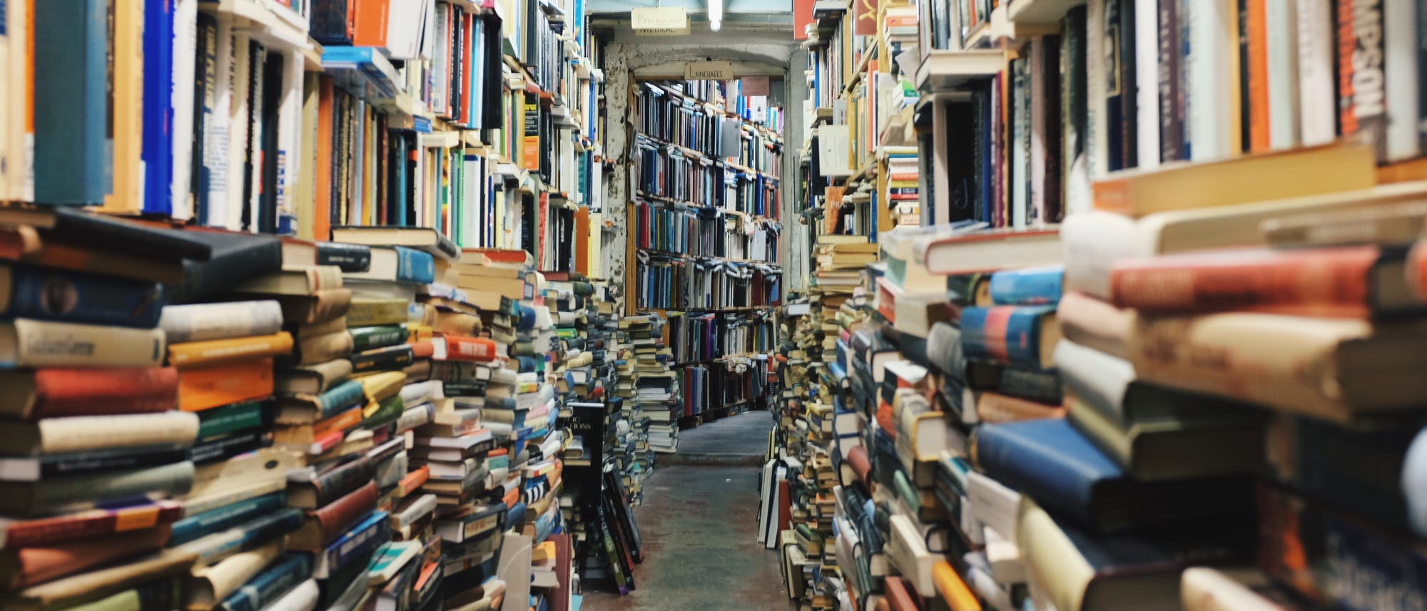enger Gang zwischen gestapelten Büchern vor vollen Bücherregalen enger Gang zwischen gestapelten Büchern vor vollen Bücherregalen