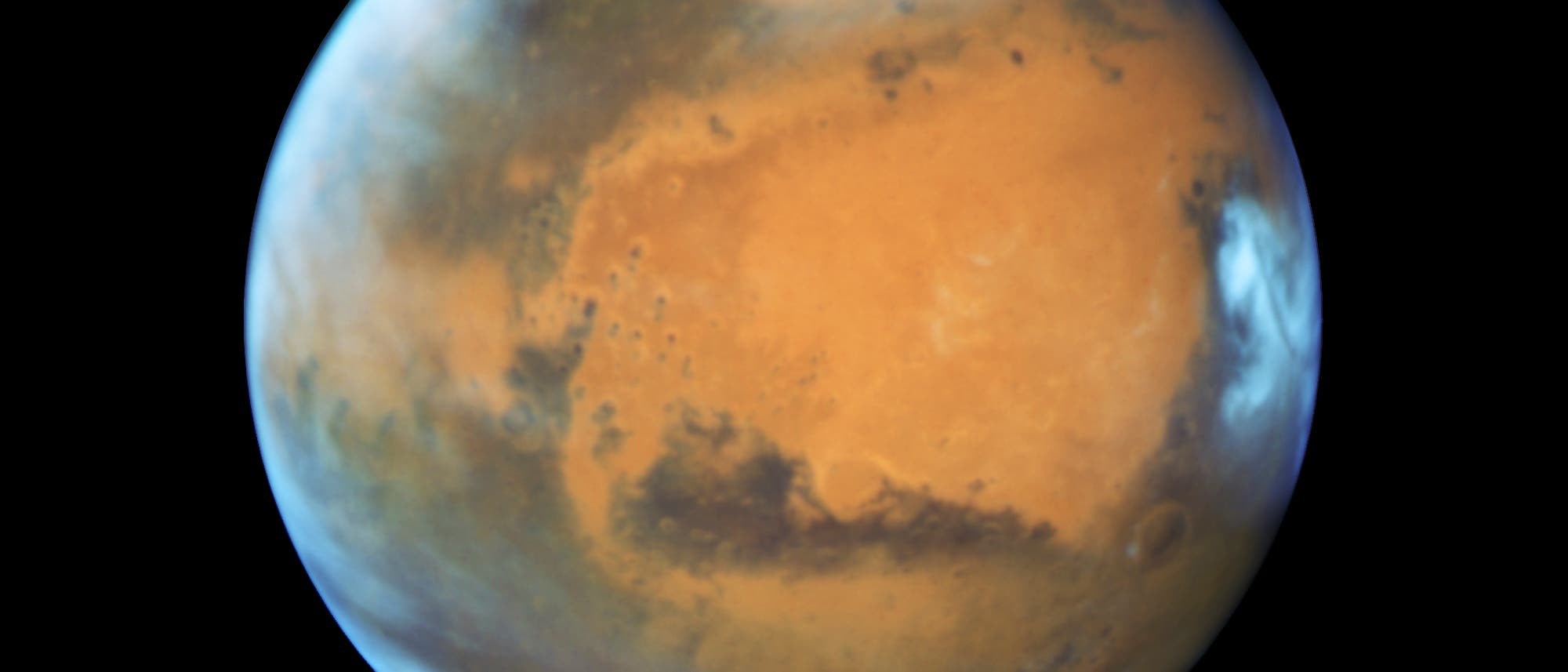 Eisbedeckte Polkappen, Wolken in der Atmosphäre, eine rostfarbene Landschaft, die von dunkleren Flecken durchsetzt ist. So präsentiert sich unser Nachbar auf diesem Bild, welches das Hubble-Weltraumteleskop am 12. Mai 2016 aufgenommen hat. Damals befand sich der Rote Planet in einer Entfernung von 50 Millionen Kilometern zur Erde. Bemerkenswert sind vor allem die Eisschilde an Nord- und Südpol: Im Norden hat sich das Eis während des gegenwärtigen Marssommers weit zurückgezogen, während es im Süden weiter ausgedehnt ist. Dafür behindern dort Wolken die Sicht. Die große dunkle Region rechts ist Syrtis Major Planitia, die zu den ersten Oberflächenformationen gehört, die auf dem Mars entdeckt und beschrieben wurden - es handelt sich um einen alten und inaktiven Vulkan. Südlich davon schließt sich die die riesige Hellas Planitia an, die vor 3,5 Milliarden Jahren durch einen Asteroideneinschlag entstand. Die orange Fläche im Zentrum wiederum ist Arabia Terra, ein ausgedehntes Hochland, das stark verkratert und erodiert ist. Eisbedeckte Polkappen, Wolken in der Atmosphäre, eine rostfarbene Landschaft, die von dunkleren Flecken durchsetzt ist. So präsentiert sich unser Nachbar auf diesem Bild, welches das Hubble-Weltraumteleskop am 12. Mai 2016 aufgenommen hat. Damals befand sich der Rote Planet in einer Entfernung von 50 Millionen Kilometern zur Erde. Bemerkenswert sind vor allem die Eisschilde an Nord- und Südpol: Im Norden hat sich das Eis während des gegenwärtigen Marssommers weit zurückgezogen, während es im Süden weiter ausgedehnt ist. Dafür behindern dort Wolken die Sicht. Die große dunkle Region rechts ist Syrtis Major Planitia, die zu den ersten Oberflächenformationen gehört, die auf dem Mars entdeckt und beschrieben wurden - es handelt sich um einen alten und inaktiven Vulkan. Südlich davon schließt sich die die riesige Hellas Planitia an, die vor 3,5 Milliarden Jahren durch einen Asteroideneinschlag entstand. Die orange Fläche im Zentrum wiederum ist Arabia Terra, ein ausgedehntes Hochland, das stark verkratert und erodiert ist.