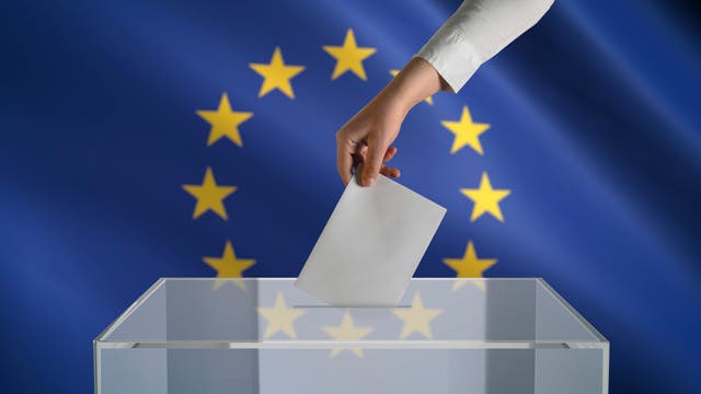 Eine Hand wirft einen Zettel in einen durchsichtigen Kasten. Im Hintergrund die Europaflagge.