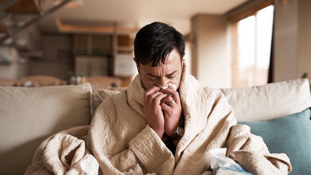 Ein Mann sitzt in eine Decke gehüllt auf der Couch und schneuzt sich die Nase.