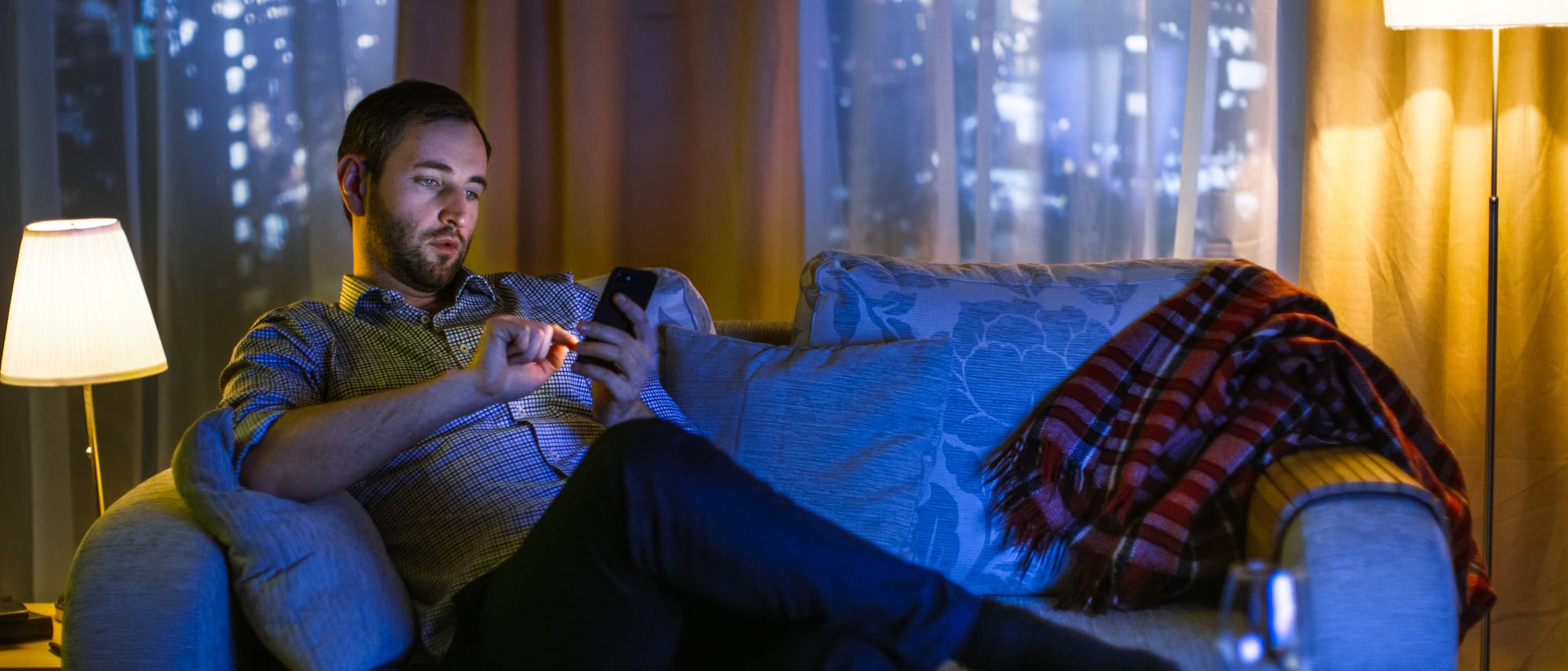 Mann sitzt mit Handy auf der Couch, im blauen Licht des Fernsehers Mann sitzt mit Handy auf der Couch, im blauen Licht des Fernsehers