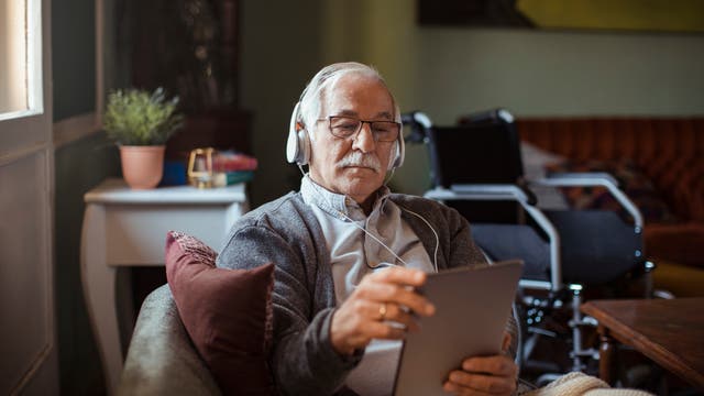 Älterer Mann mit Kopfhörern sitzt in einem Sessel und benutzt ein Tablet.