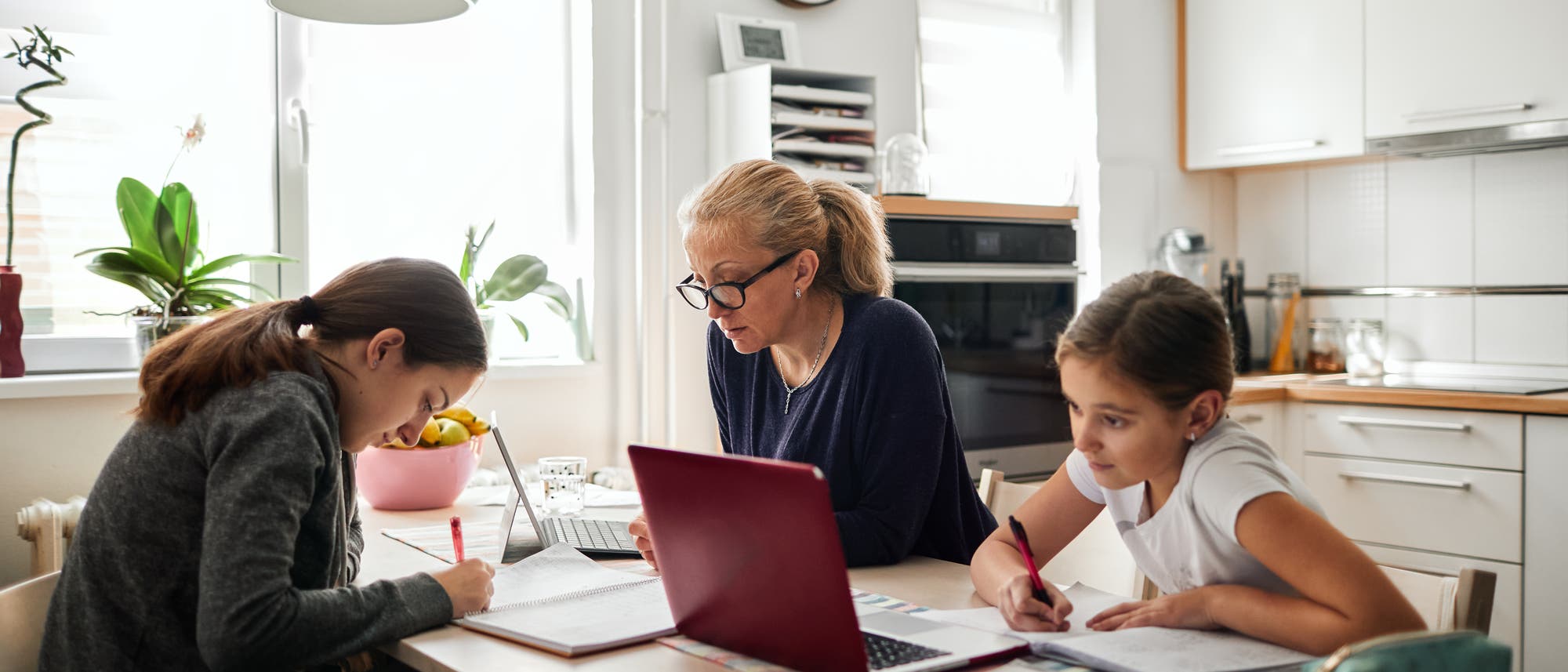 Eine Mutter sitzt mit ihren zwei Töchtern über Heften und Arbeitsblättern. Eine Mutter sitzt mit ihren zwei Töchtern über Heften und Arbeitsblättern.