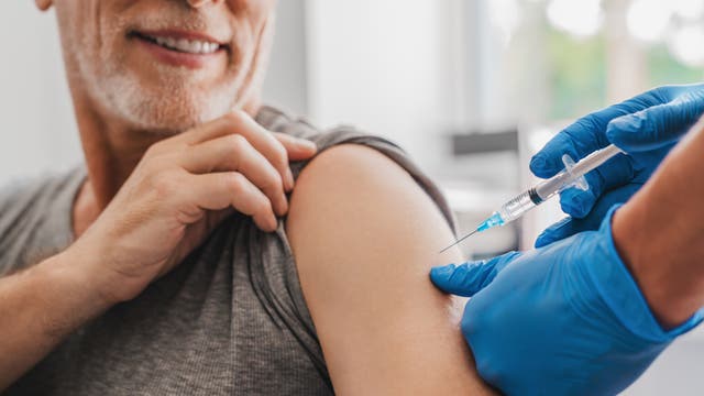 Älterer Herr mit Vollbart bekommt eine Injektion in den Arm. Das Foto ist vermutlich auch nicht echt, aber hier ist immerhin Flüssigkeit in der Spritze. Das ist nicht bei allen Impfbildern so. 
