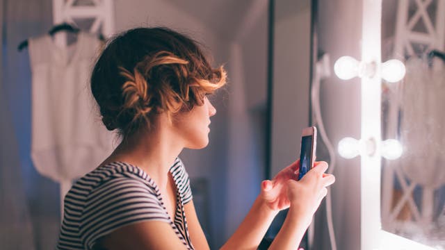 Frau vor beleuchtetem Spiegel mit Smartphone in den Händen