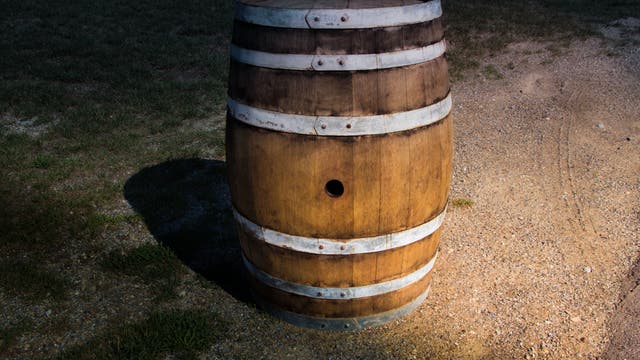 Ein leeres Weinfass mit offenem Spundloch steht halb beleuchtet auf sandigem Boden.