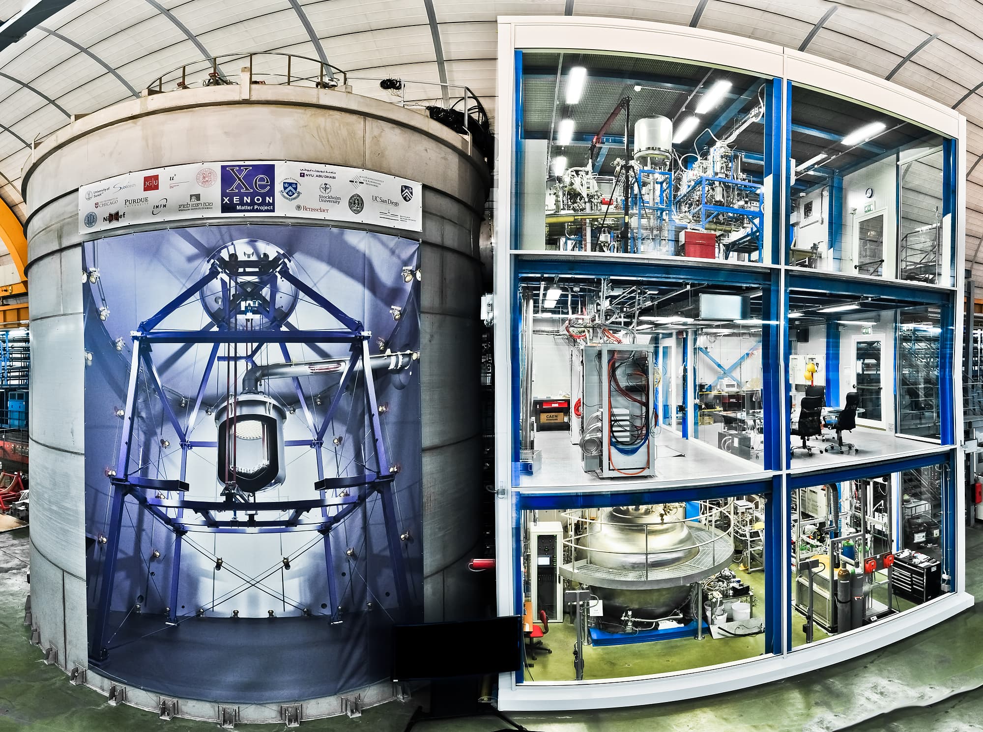 Wassertank und Gerätehaus von XENON1T – auf dem Tank ist eine Innenansicht des Tanks mit dem in der Mitte aufgehängten Detektor aufgedruckt. Wassertank und Gerätehaus von XENON1T – auf dem Tank ist eine Innenansicht des Tanks mit dem in der Mitte aufgehängten Detektor aufgedruckt.