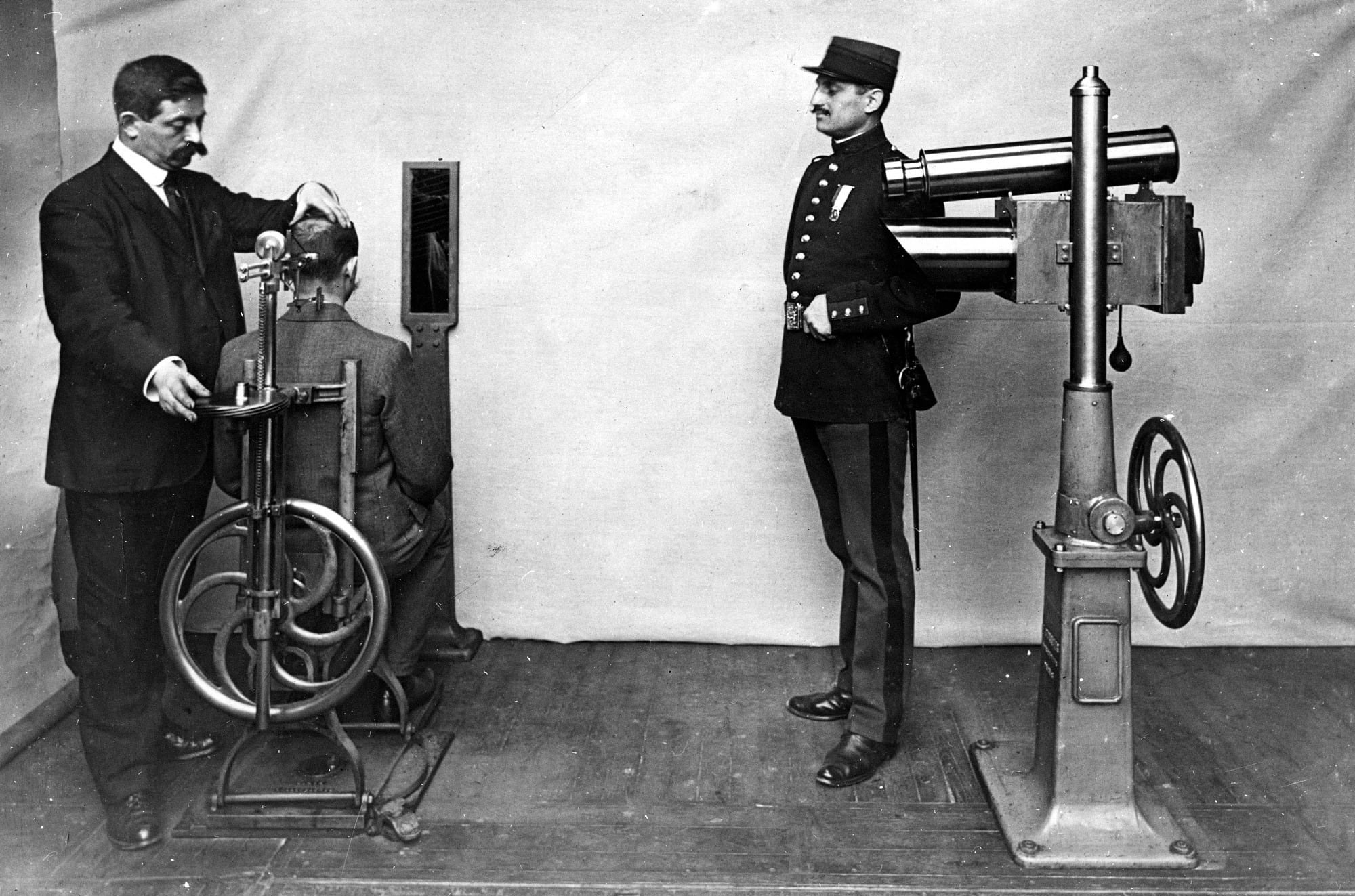 Zwei Personen, eine davon in Uniform, vermessen einen sitzenden Mann Zwei Personen, eine davon in Uniform, vermessen einen sitzenden Mann