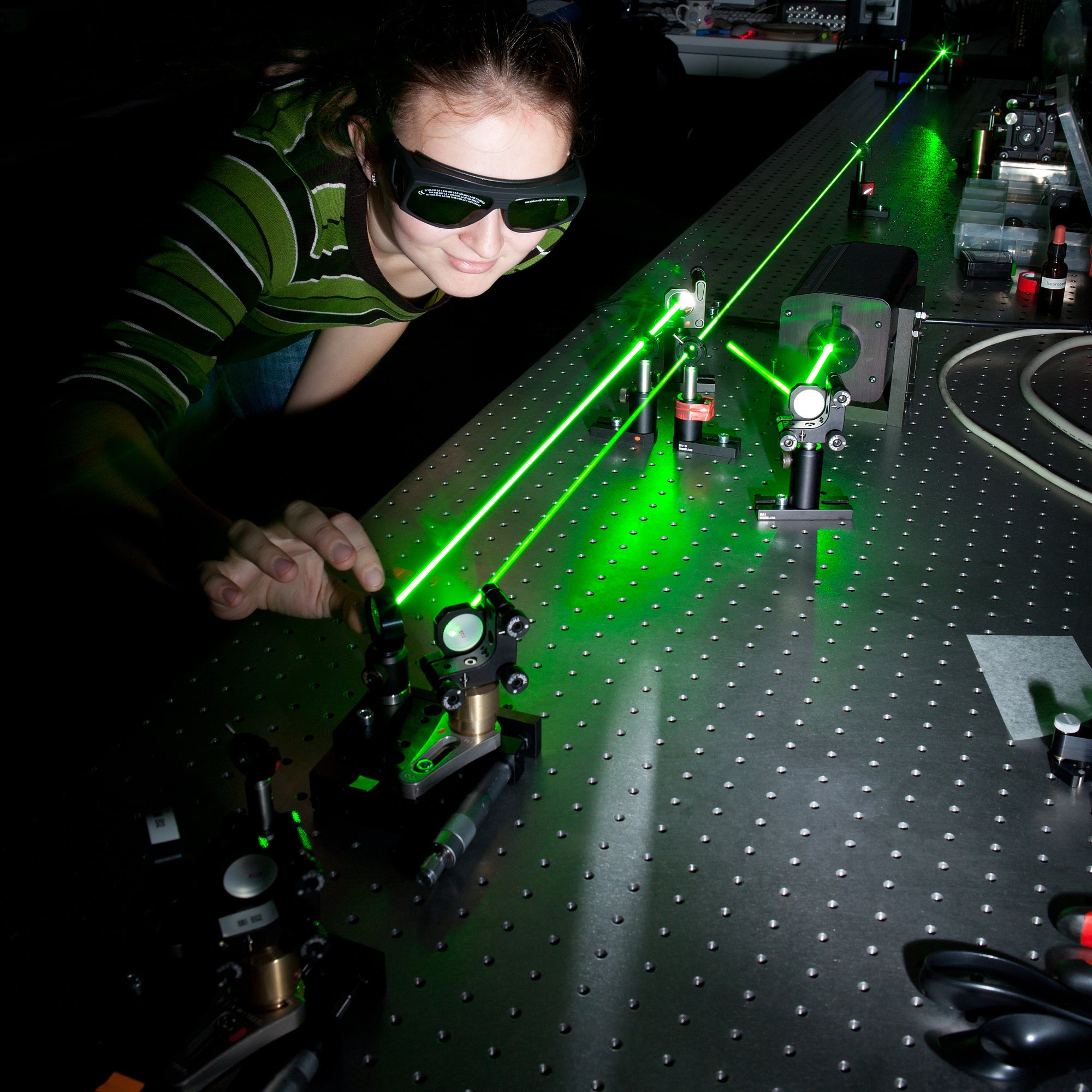 Eine Forscherin an einem Versuchsaufbau mit grünen Lasern, Strahlteilern und Spiegeln. Eine Forscherin an einem Versuchsaufbau mit grünen Lasern, Strahlteilern und Spiegeln.